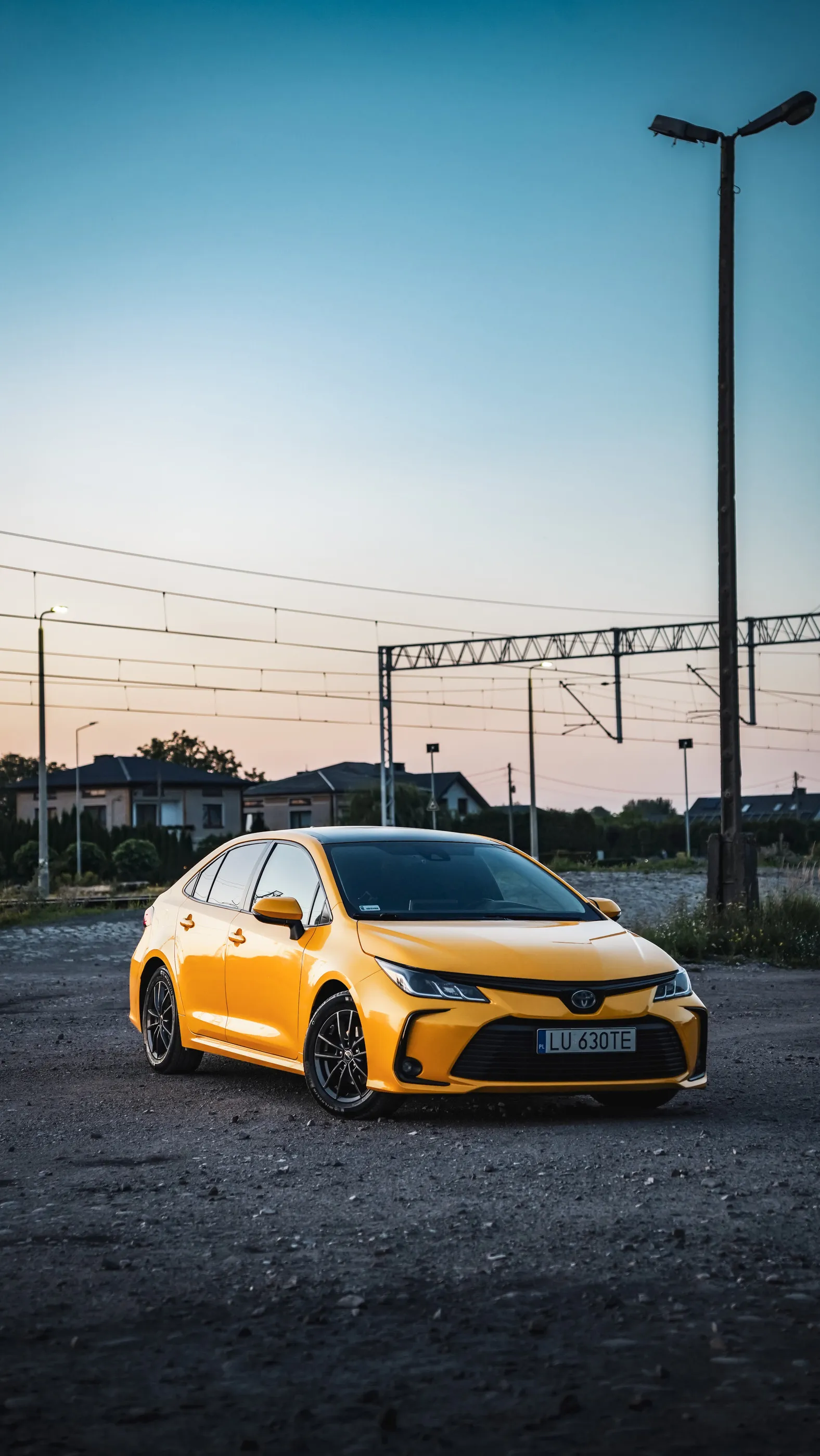 Żółta Toyota Corolla na placu o zmierzchu, w tle sieć trakcyjna